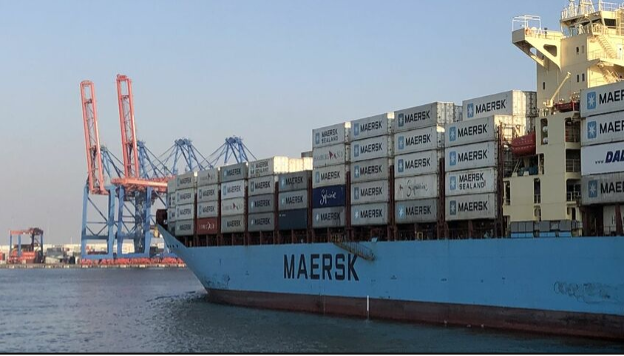 A Maersk container ship docked at a port, loaded with stacked containers, alongside gantry cranes